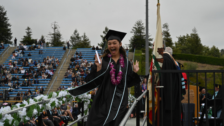 Student dressed in black graduation cap and gown celebrates as she walks off stage with diploma